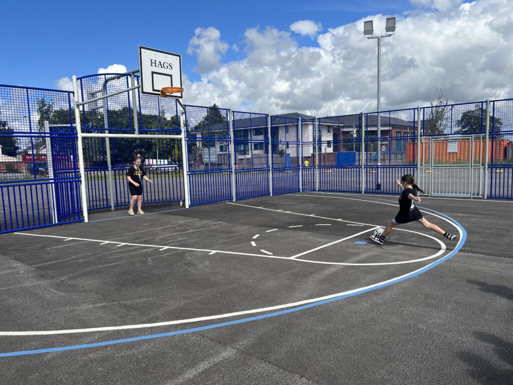 An action shot of two members of the Youth Forum at Alder Hey playing football on the multi use games area. One young person is in goal and the other is about to strike the ball.