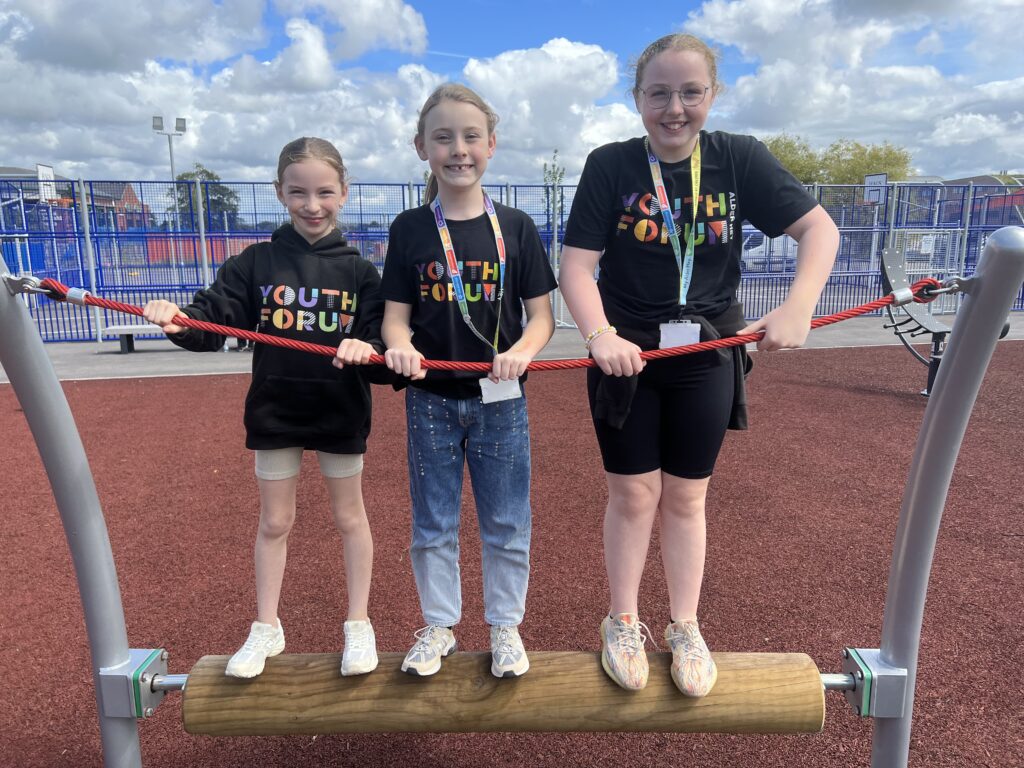 Three Youth Forum at Alder Hey members stand smiling on a piece of trim trail equipment.