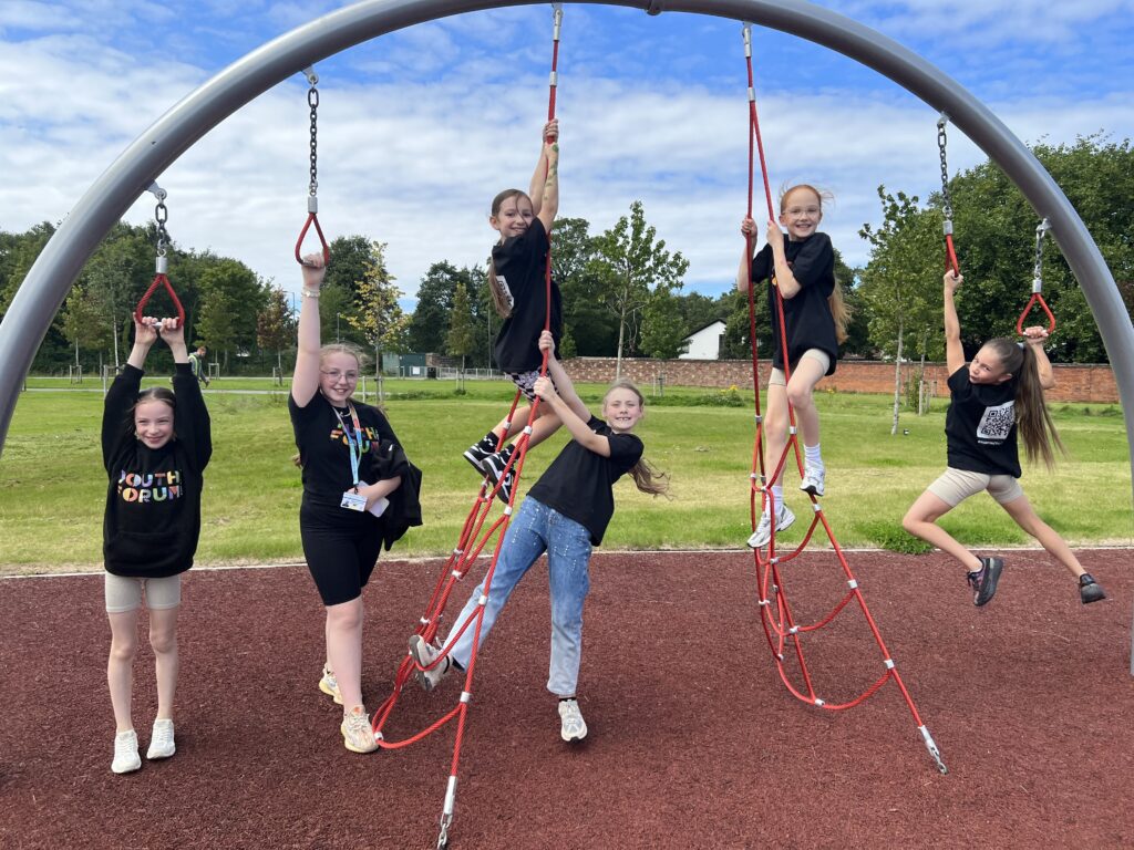 Six members of the Youth Forum at Alder Hey are smiling as they hang on to a piece of trim trail equipment.