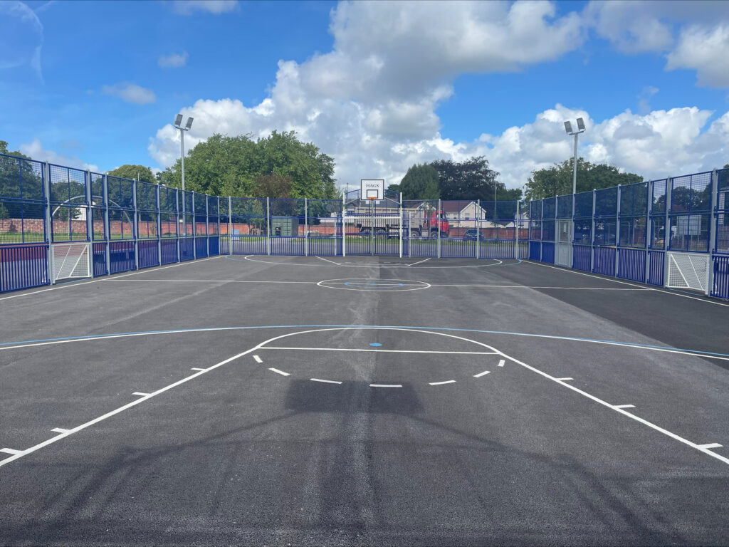 A wider view of the multi use games area on Springfield park showing a basketball hoop and goal, as well as the overhead lighting and side fencing.