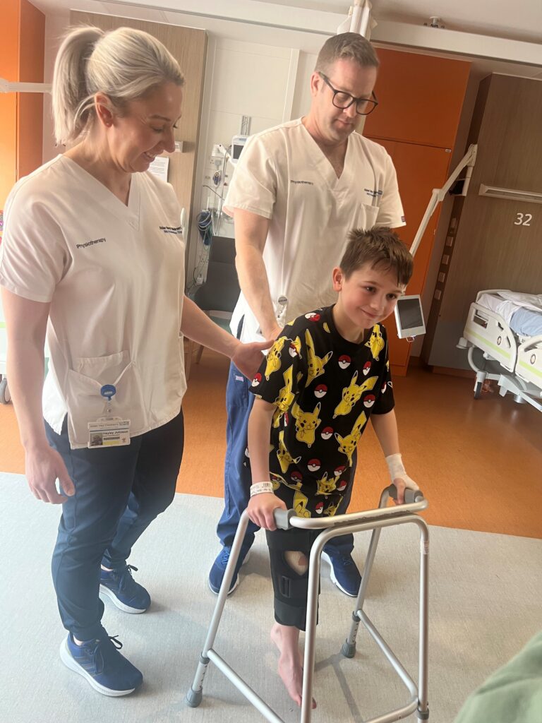 Alfie walking with his zimmerframe with two Alder Hey Physiotherapists