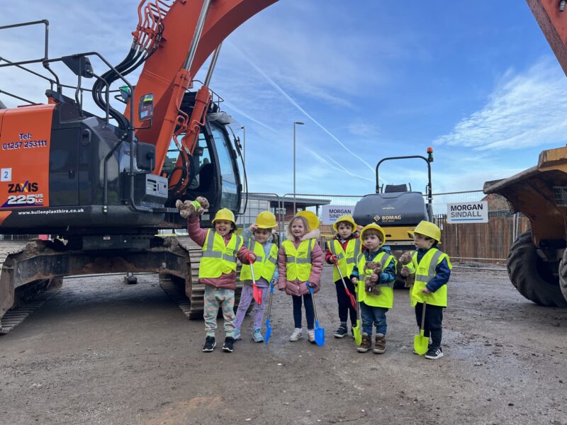 Members of the Alder Hey Nursery post next to a digger holding toy spades and wearing PPE.