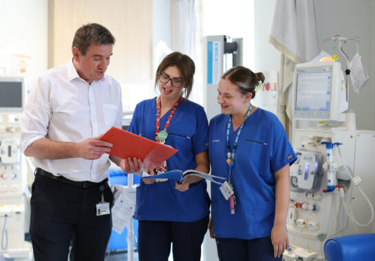 Photo of two nurses looking at a red clipboard being held by a man in a white shirt