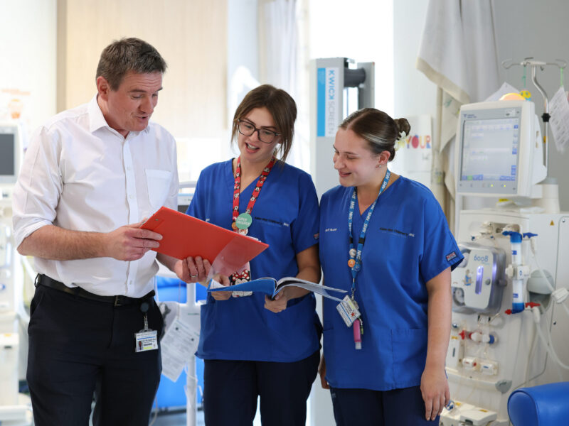 Photo of two nurses looking at a red clipboard being held by a man in a white shirt
