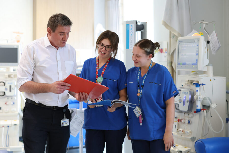 Photo of two nurses looking at a red clipboard being held by a man in a white shirt