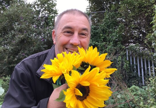 A member of the Alder Hey FRESH CAMHS team smiling outdoors while holding bright yellow sunflowers.