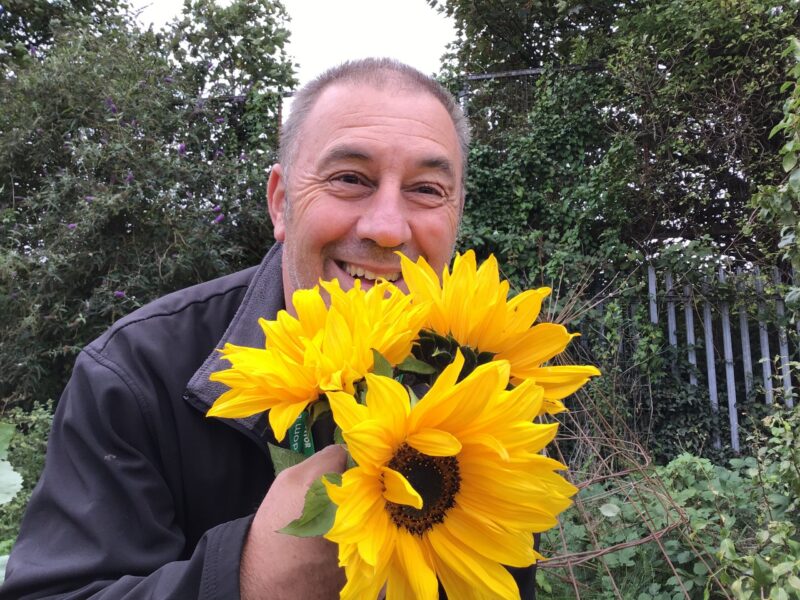 A member of the Alder Hey FRESH CAMHS team smiling outdoors while holding bright yellow sunflowers.