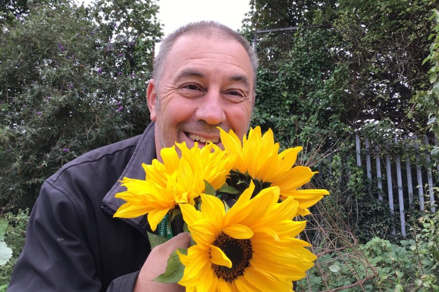 A member of the Alder Hey FRESH CAMHS team smiling outdoors while holding bright yellow sunflowers.