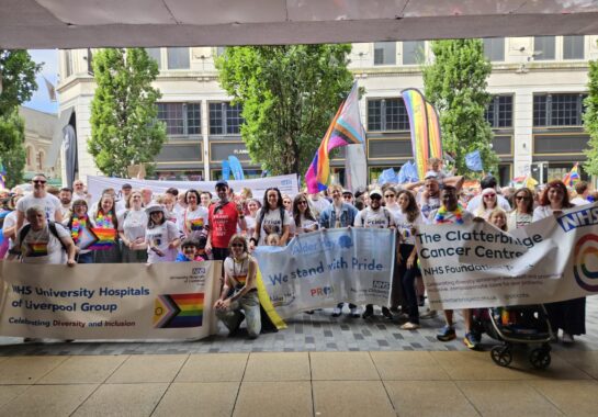 Photo of many people at Liverpool Pride. There are many banners which show support for Liverpool Pride such as One NHS