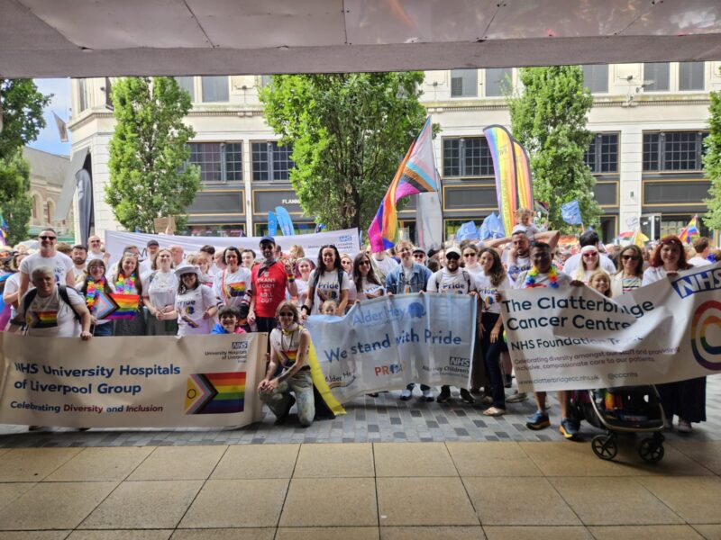 Photo of many people at Liverpool Pride. There are many banners which show support for Liverpool Pride such as One NHS