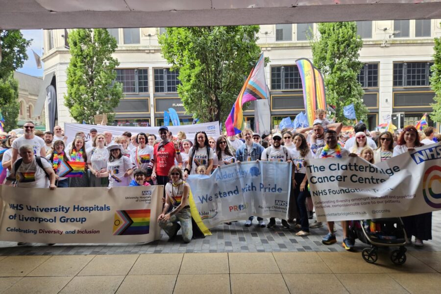 Photo of many people at Liverpool Pride. There are many banners which show support for Liverpool Pride such as One NHS
