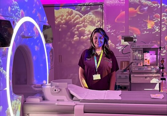 Radiographer Kirsty Hillis stands beside an MRI scanner in a child-friendly room with colourful lighting and underwater projections.