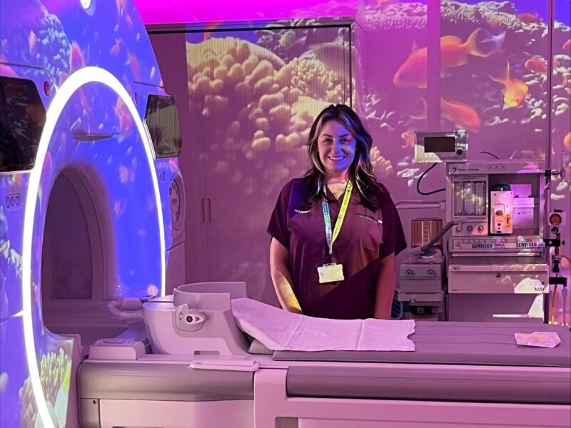 Radiographer Kirsty Hillis stands beside an MRI scanner in a child-friendly room with colourful lighting and underwater projections.