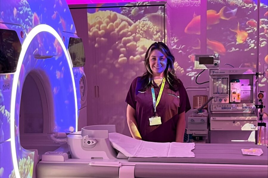 Radiographer Kirsty Hillis stands beside an MRI scanner in a child-friendly room with colourful lighting and underwater projections.